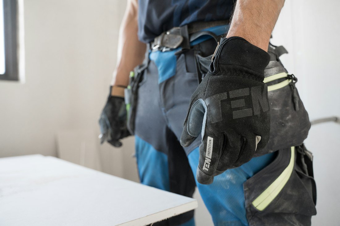 Tradesman in work gloves and a tool belt beside a sheet of drywall