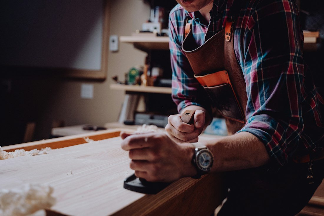 Carpenter in a plaid shirt and leather apron planing a board